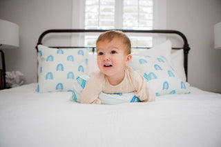 A baby with light brown hair and a beige outfit lies on white bed sheets featuring a Toddler Sleeping Bundle: Organic Pillow + Blue Rainbows Case. The room is brightly lit by natural sunlight streaming through the window in the background.
