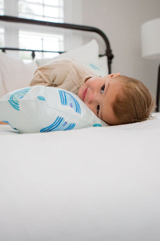A child rests on a white bed, their head on a pillow from the Toddler Sleeping Bundle: Organic Pillow + Blue Rainbows Case. The room features muted colors, a window in the background, and a modern floor lamp beside the bed.