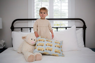 A toddler in beige attire stands on a bed with pristine white linens, next to a large plush sheep and an organic, machine washable Toddler Sleeping Pillow Case showcasing a yellow school bus pattern. The bed is set against a window with shutters.