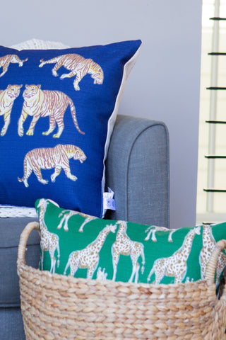 A blue throw pillow with a tiger print lies on a gray sofa alongside a white knitted blanket. The Giraffes on Grass Lumbar Pillow adds an exotic touch from its position in the woven basket on the floor, with vertical blinds partially visible in the background.