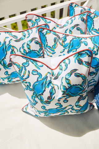 The Crab Craze pillow in shades of blue with red piping is stacked on a light-colored outdoor bench, set against green foliage.