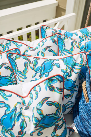 The vibrant Crab Craze, Shades of Blue Outdoor Pillow with red piping is stacked on a white bench in a sunlit porch or patio, accompanied by a blue woven bag, showcasing the durability of SDS Shield® Outdoor Fabric.