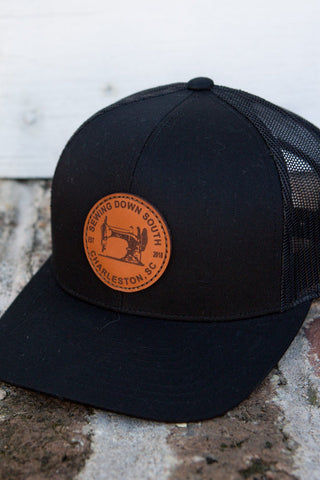 The Sewing Down South Leather Patch Trucker, Black on Black, features a vintage logo on a brown leather patch and sits atop a brick surface near a white wooden wall and iron railing.