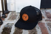 The Sewing Down South Leather Patch Trucker, Black on Black, features a vintage logo on a brown leather patch and sits atop a brick surface near a white wooden wall and iron railing.