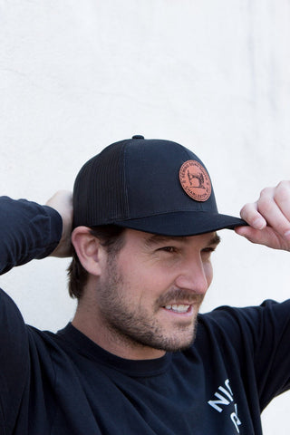 A man adjusts his Sewing Down South Leather Patch Trucker, Black on Black, while wearing a black long-sleeve shirt and standing in front of a light-colored wall.