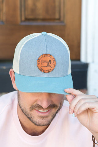 A man sits on brick steps wearing a grey Sewing Down South Leather Patch Trucker hat with a vintage logo, paired with a light pink sweatshirt and bracelet. He slightly tilts his head and touches the caps brim, framed by a wooden door and white wall in the background.