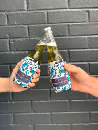 Two hands clink beer bottles in vibrant blue and white Crab Craze Koozie neoprene covers, set against a gray brick wall—perfect for sharing from the Crab Craze Koozie, 4 PACK (Skinny or Standard).