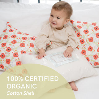 A toddler, dressed in beige and smiling brightly, sits on a bed with floral pillows while holding the Toddler Sleeping Bundle: Organic Pillow + Blue Rainbows Case. The plastic-wrapped pillows label reads 100% GOTS Certified Organic Cotton Shell, highlighting its eco-friendly and hypoallergenic features.