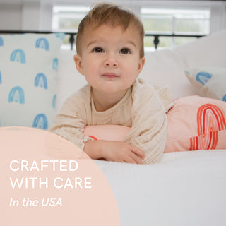 A toddler with light brown hair rests on a bed with a Toddler Sleeping Bundle: Organic Pillow + Blue Rainbows Case. The child wears a light beige outfit. Text in the bottom left reads, Crafted with care in the USA using GOTS Certified Organic Cotton.
