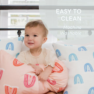 A baby sits among pillows with rainbow patterns, featuring the Toddler Sleeping Bundle: Organic Pillow + Blue Rainbows Case. A translucent circle reads Easy to Clean, Machine Washable. In the softly lit room, a window in the background envelops everything in a gentle glow.