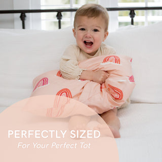 A toddler smiles on a white couch while hugging an organic pillow with a colorful blue rainbows case. Text reads, Perfectly Sized For Your Perfect Tot. Product: Toddler Sleeping Bundle: Organic Pillow + Blue Rainbows Case.