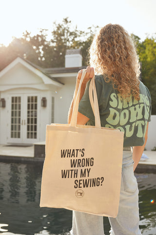 A person with curly hair and a green text t-shirt stands by a pool holding the Whats Wrong with My Sewing Tote, a 100% cotton beige bag featuring an expandable bottom. The scene is sunlit, with a house in the background.