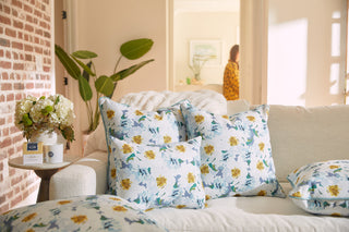 A cozy living room features a white couch with Sewing Down Souths Blossoming Peonies Lumbar Pillow in a yellow and blue floral design. A side table holds a vase of white roses, while someone stands near the doorway.