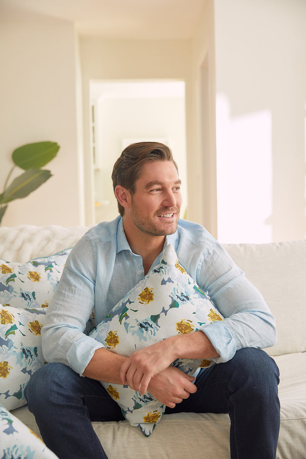 A man in a light blue shirt relaxes on a white couch, holding a floral pillow with yellow flowers. He gazes aside with a soft smile as natural light and greenery fill the room. The Bee & Florals Collection: Blossoming Peonies Lumbar Pillow beautifully complements the bright scene.