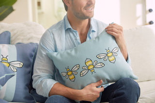 A man in a light blue shirt holds a Bee & Florals Collection: Bee Trio Lumbar Pillow by Sewing Down South. He sits on the sofa with another similar pillow nearby, in a softly lit, cozy room that exudes warmth and charm.