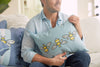A man in a light blue shirt holds a Bee & Florals Collection: Bee Trio Lumbar Pillow by Sewing Down South. He sits on the sofa with another similar pillow nearby, in a softly lit, cozy room that exudes warmth and charm.