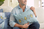 A man in a light blue shirt holds a Bee & Florals Collection: Bee Trio Lumbar Pillow by Sewing Down South. He sits on the sofa with another similar pillow nearby, in a softly lit, cozy room that exudes warmth and charm.