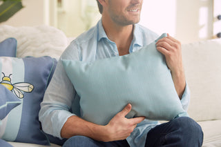 A person in a light blue shirt sits on a sofa with a Bee & Florals Collection: Bee Trio Lumbar Pillow. The calm, cozy setting with soft lighting enhances the scenes charm. This USA-crafted pillow by Sewing Down South adds warmth to the ambiance.