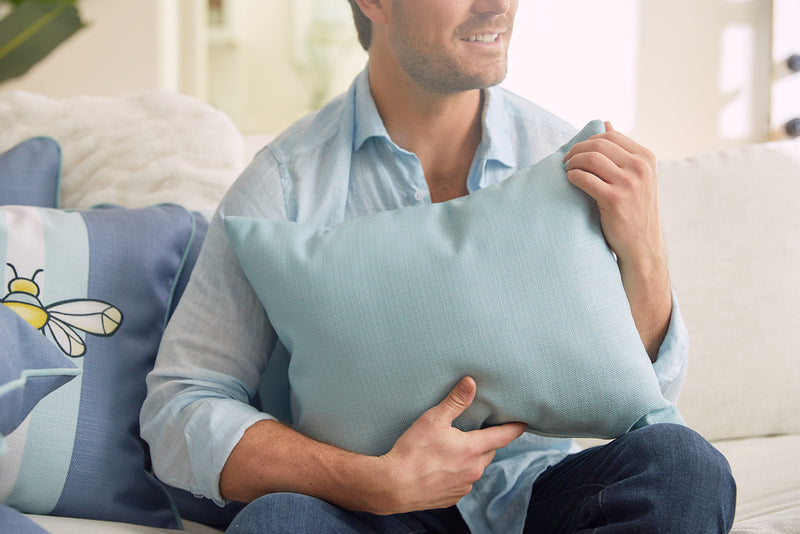 A person in a light blue shirt sits on a sofa with a Bee & Florals Collection: Bee Trio Lumbar Pillow. The calm, cozy setting with soft lighting enhances the scenes charm. This USA-crafted pillow by Sewing Down South adds warmth to the ambiance.