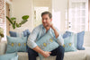 A man sits on a sofa in a brightly lit living room, surrounded by Bee & Florals Collection: Bee Trio Lumbar Pillows, proudly made in the USA. In his light blue shirt and dark pants, he relaxes amidst plants, a brick wall, and white doors.