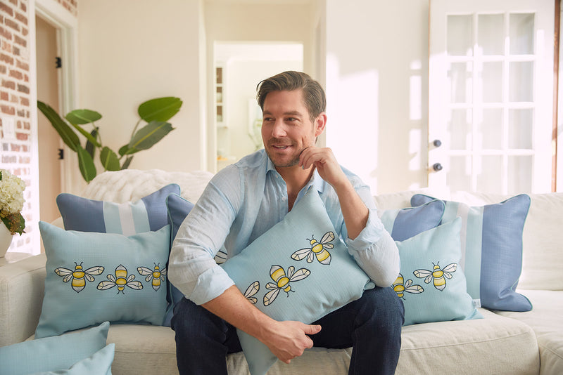 A man sits on a sofa in a brightly lit living room, surrounded by Bee & Florals Collection: Bee Trio Lumbar Pillows, proudly made in the USA. In his light blue shirt and dark pants, he relaxes amidst plants, a brick wall, and white doors.