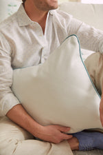 A person in a light-colored, Victorian floral-patterned shirt sits on a beige couch, holding the Bee & Florals Collection: Victorian Floral in Blue Pillow with teal piping. Their head is turned away. Light brown pants and blue shoes complete the look.