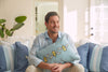 A man smiles on a sofa, wearing a light blue shirt, holding the Bee Trio Lumbar Pillow from the Bee & Florals Collection by Sewing Down South. Sunlight highlights its decorative bee designs and perfectly matches the surrounding blue and white striped pillows.