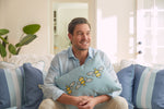 A man smiles on a sofa, wearing a light blue shirt, holding the Bee Trio Lumbar Pillow from the Bee & Florals Collection by Sewing Down South. Sunlight highlights its decorative bee designs and perfectly matches the surrounding blue and white striped pillows.