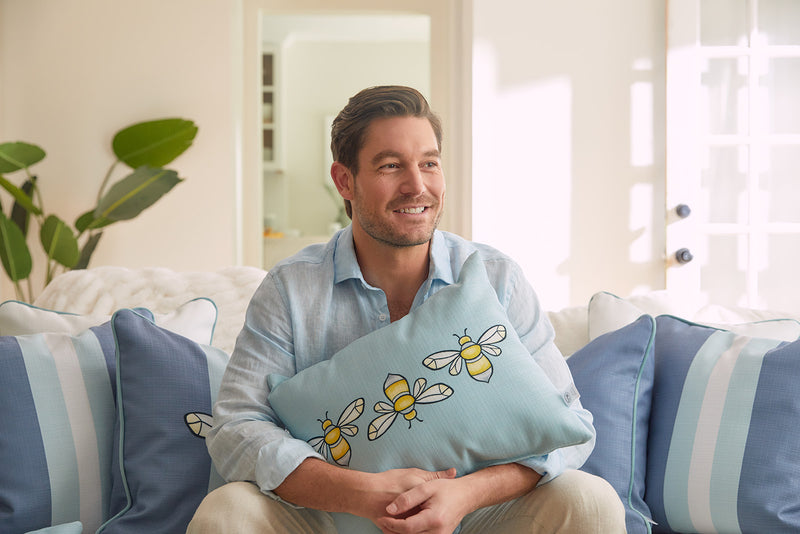 A man smiles on a sofa, wearing a light blue shirt, holding the Bee Trio Lumbar Pillow from the Bee & Florals Collection by Sewing Down South. Sunlight highlights its decorative bee designs and perfectly matches the surrounding blue and white striped pillows.