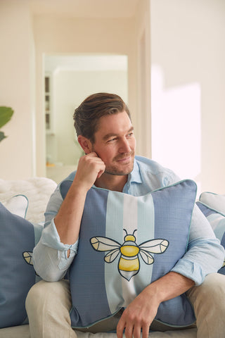 A man with short hair and a beard relaxes on a couch, resting his head on a Bee & Florals Collection: Bee Stripe Pillow from Sewing Down South. In a light blue shirt, he gazes thoughtfully into the distance.