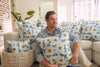 A person sits on a couch, holding a Blossoming Peonies Pillow from the Bee & Florals Collection. The sofa is decorated with complementary blue and yellow floral pillows by Sewing Down South®. A woven basket and potted plant enhance the bright, sunlit rooms charm.