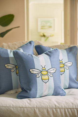 Bee & Florals Collection: Bee Stripe Pillows in blue and white are displayed on a beige sofa, with a plant, blurred picture, and flowers on a shelf in the background, creating a cozy atmosphere.