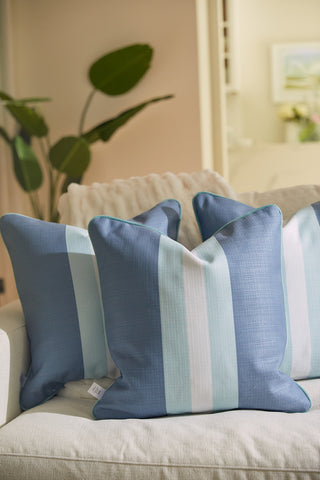 A cozy living room showcases a cream sofa with three Bee & Florals Collection: Bee Stripe Pillows featuring light blue and white stripes. A large green plant enhances the inviting atmosphere, adding a natural touch in the background.
