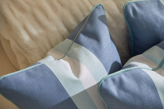 Close-up of Bee & Florals Collection: Bee Stripe Pillow, showcasing a blue and white checkered design with light blue piping. It offers a soft look against textured beige fabric, contrasting strikingly with a wavy patterned backdrop.