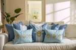 A cozy beige sofa from Sewing Down South is decorated with blue and white throw pillows, featuring the whimsical Bee & Florals Collection: Bee Trio Lumbar Pillow. Sunlight creates soft shadows as a plant adds greenery to the serene scene. Made in the USA.
