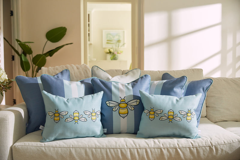 A cozy beige sofa from Sewing Down South is decorated with blue and white throw pillows, featuring the whimsical Bee & Florals Collection: Bee Trio Lumbar Pillow. Sunlight creates soft shadows as a plant adds greenery to the serene scene. Made in the USA.