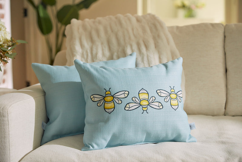 The USA-made Bee Trio Lumbar Pillow from Sewing Down Souths Bee & Florals Collection, in light blue, adorns a cream sofa. Embroidered bees with yellow and black stripes and white wings add charm against a backdrop of green plants and a blurred floral arrangement.