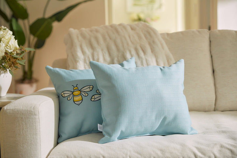 A cozy living room features a beige sofa with light blue cushions, including the Bee & Florals Collection: Bee Trio Lumbar Pillow, made in the USA. Its cartoon bee design pairs well with a draped fluffy throw and vibrant plant in the background.