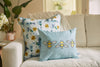Two decorative pillows sit on the cream sofa. One flaunts a floral design in blue and yellow, while the Bee & Florals Collection: Bee Trio Lumbar Pillow, crafted in the USA by Sewing Down South, features three delightful bees on light blue. A plant and flowers provide a backdrop.