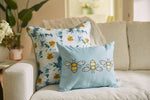 Two decorative pillows sit on the cream sofa. One flaunts a floral design in blue and yellow, while the Bee & Florals Collection: Bee Trio Lumbar Pillow, crafted in the USA by Sewing Down South, features three delightful bees on light blue. A plant and flowers provide a backdrop.