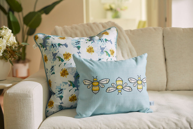 Two decorative pillows sit on the cream sofa. One flaunts a floral design in blue and yellow, while the Bee & Florals Collection: Bee Trio Lumbar Pillow, crafted in the USA by Sewing Down South, features three delightful bees on light blue. A plant and flowers provide a backdrop.