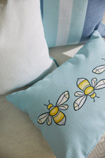 Close-up of decorative pillows on a light surface. The Bee & Florals Collection: Bee Trio Lumbar Pillow from Sewing Down South features light blue fabric with yellow and white bee patterns. Another pillow has vertical blue and white stripes, while a solid white one completes the set. Made in the USA.