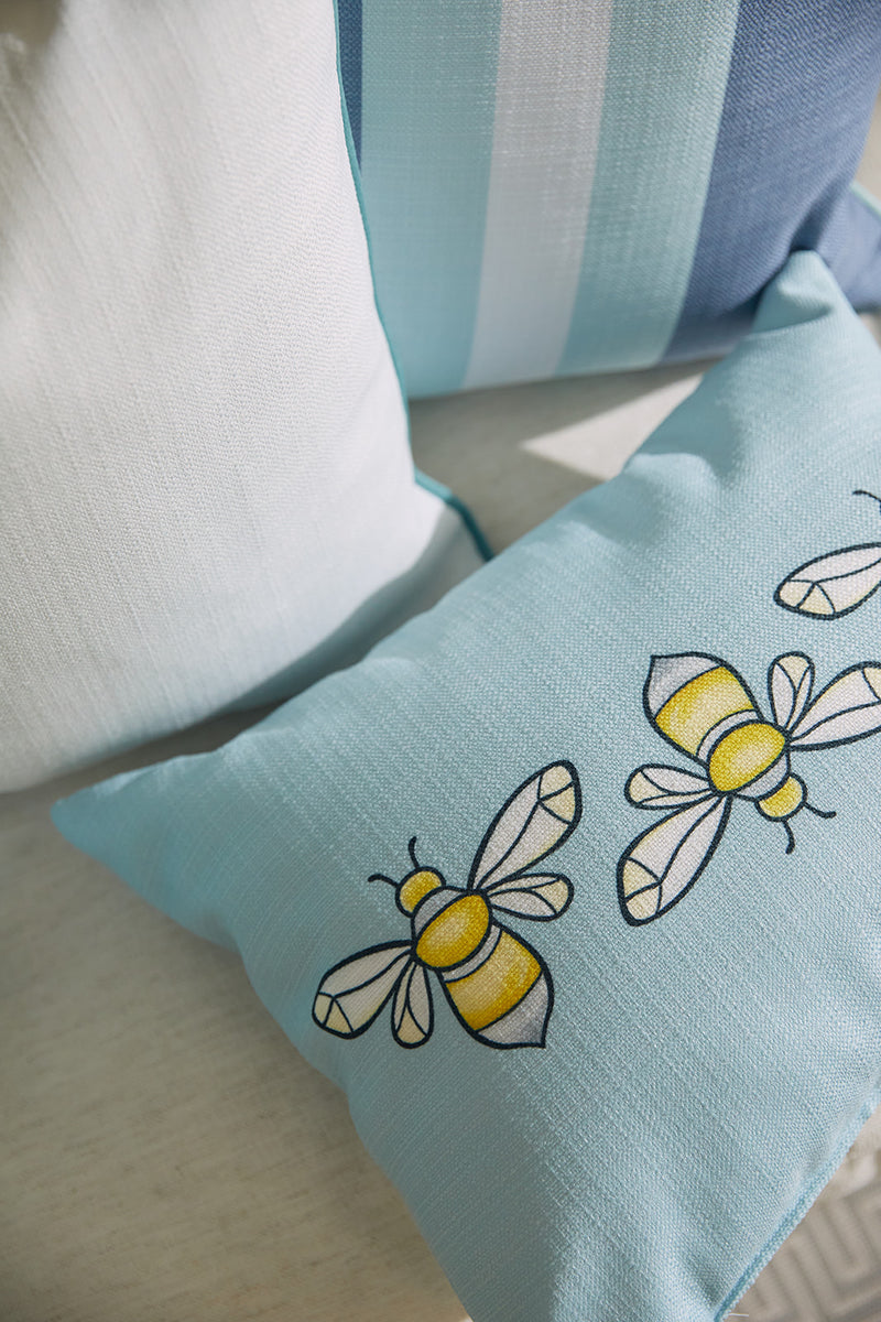 Close-up of decorative pillows on a light surface. The Bee & Florals Collection: Bee Trio Lumbar Pillow from Sewing Down South features light blue fabric with yellow and white bee patterns. Another pillow has vertical blue and white stripes, while a solid white one completes the set. Made in the USA.