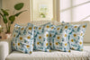 Four floral-patterned pillows, including the Bee & Florals Collection: Blossoming Peonies Pillow, are neatly arranged on a light sofa. Soft sunlight filters through a window, casting gentle shadows. A large green plant and artwork complete the serene and airy room.