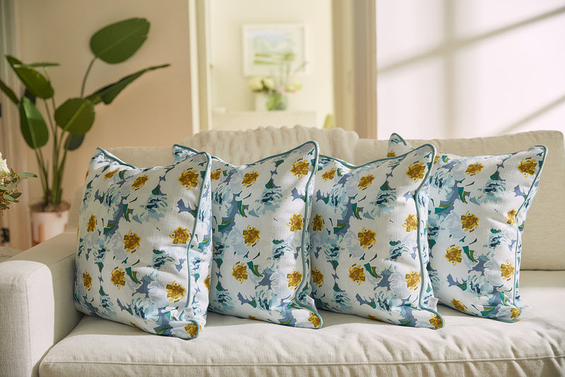 Four floral-patterned pillows, including the Bee & Florals Collection: Blossoming Peonies Pillow, are neatly arranged on a light sofa. Soft sunlight filters through a window, casting gentle shadows. A large green plant and artwork complete the serene and airy room.