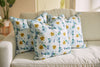 Three pillows from the Bee & Florals Collection: Blossoming Peonies Pillow by Sewing Down South®, featuring yellow and blue flowers, are arranged on a light sofa with a blurred plant and part of the room in the background.
