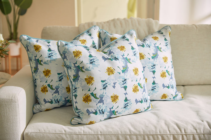 Three pillows from the Bee & Florals Collection: Blossoming Peonies Pillow by Sewing Down South®, featuring yellow and blue flowers, are arranged on a light sofa with a blurred plant and part of the room in the background.