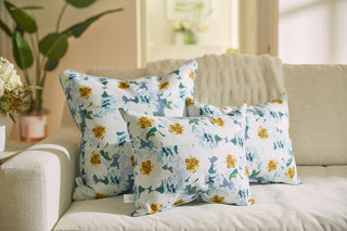 Three floral cushions, including a Blossoming Peonies Lumbar Pillow from the Bee & Florals Collection with blue and yellow flowers, are on a light sofa. A textured throw is draped over it, with a plant and bouquet in the softly lit background.