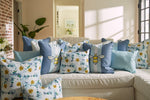 A cozy living room features a white couch with blue and white cushions, including the Bee & Florals Collection: Blossoming Peonies Pillow from Sewing Down South®, displaying delightful bee and floral designs. Sunlight streams in, casting soft shadows, while a hallway with plants and a picture frame is visible.