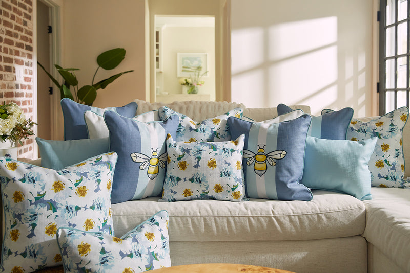 A cozy living room features a white couch with blue and white cushions, including the Bee & Florals Collection: Blossoming Peonies Pillow from Sewing Down South®, displaying delightful bee and floral designs. Sunlight streams in, casting soft shadows, while a hallway with plants and a picture frame is visible.